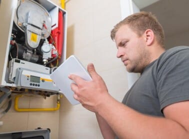 A man in a gray t-shirt is using RepairSense on his tablet as he examines the open, wall-mounted boiler. Exposed internal components and yellow pipes are visible, showcasing the intricate details he's navigating with ease. ©Mobysoft