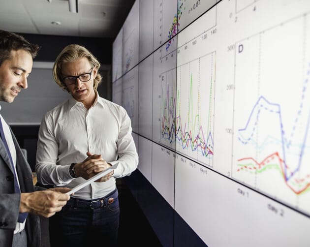 Two men in business attire are analyzing data on large screens displaying various graphs and charts. One holds a tablet, while the other gestures towards the screen. They appear focused and engaged in a modern office setting. ©Mobysoft