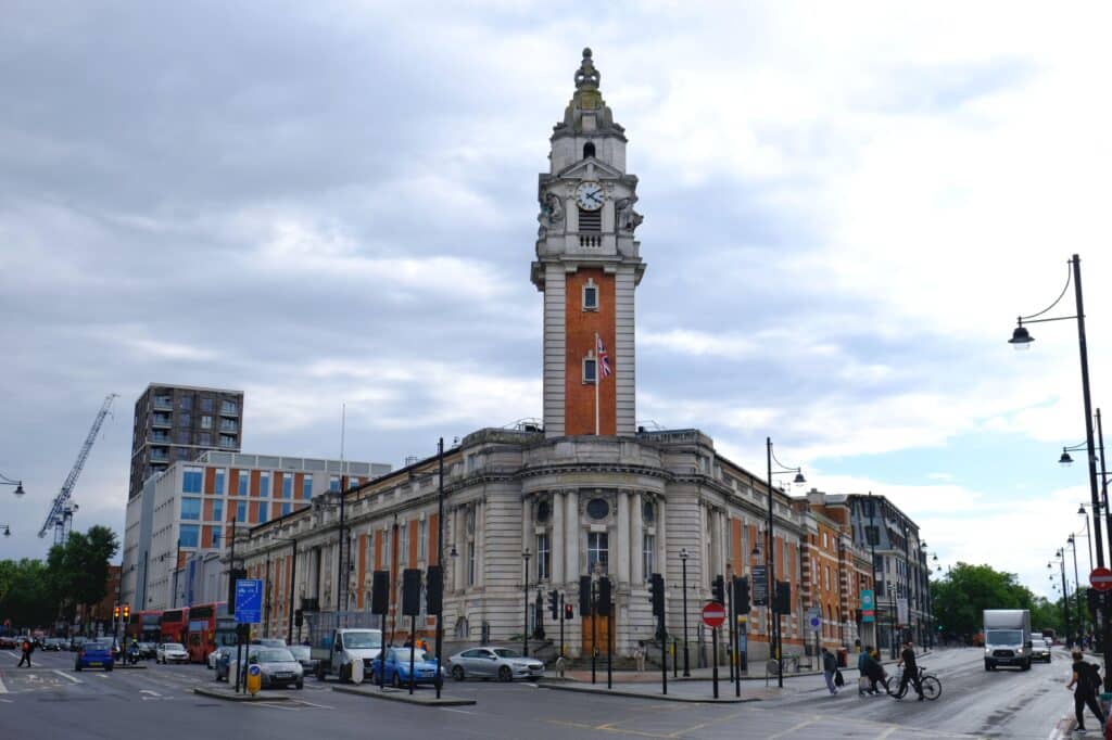 A historical building with a tall clock tower stands at an intersection, showcasing intelligent automation through its synchronized chimes. The structure features orange and white accents. People and vehicles fill the street below, while the sky remains cloudy. ©Mobysoft