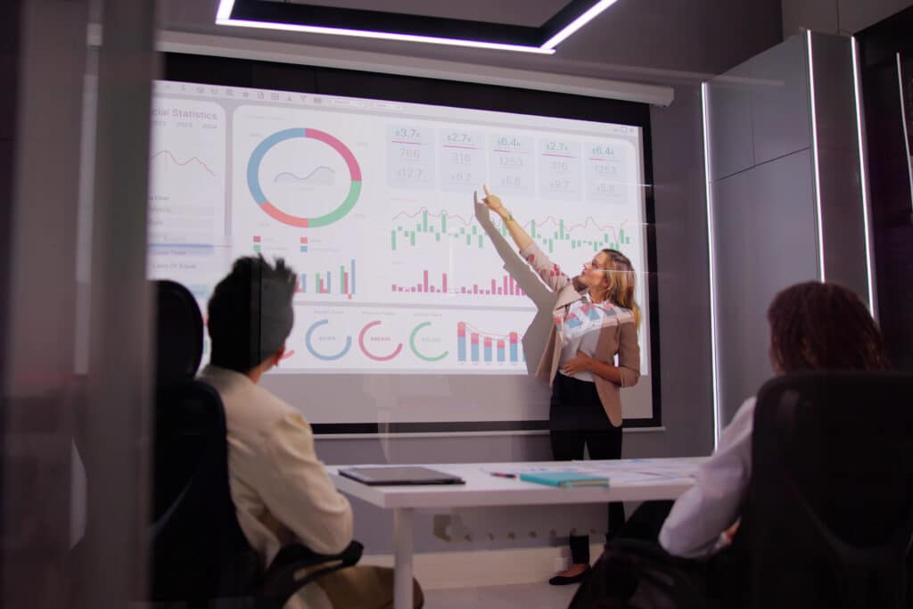 A woman stands in front of a large screen displaying graphs and charts, skillfully highlighting the data strategy. Three people sit at a table, attentively watching the presentation in the modern conference room. ©Mobysoft