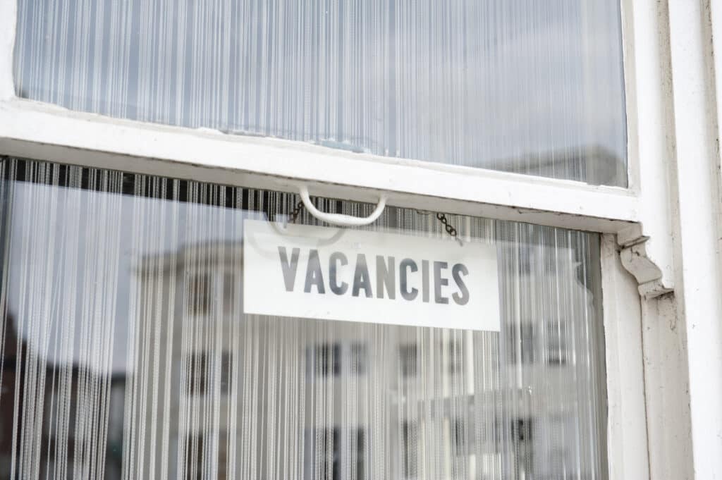 A white sign with the word "VACANCIES" in bold, black letters hangs inside a window. The window reflects blurred buildings and a cloudy sky outside. ©Mobysoft