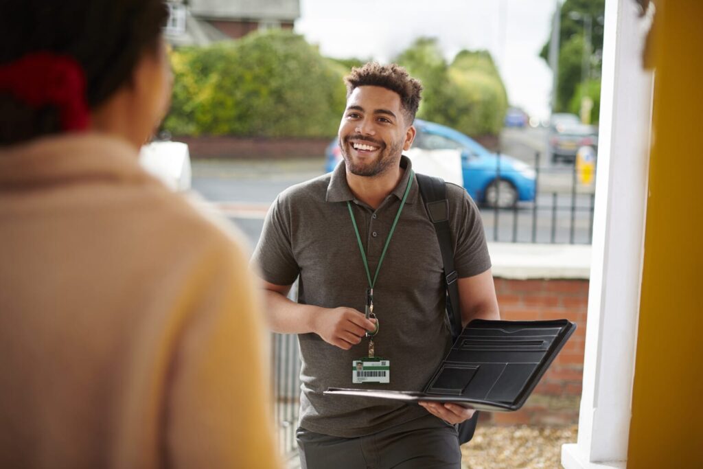 A smiling man with a laptop, ID badge, and backpack stands at a doorstep, actively engaging in resident engagement with another person. Surrounded by greenery and a street in the background, he embodies approachable professionalism outdoors. ©Mobysoft