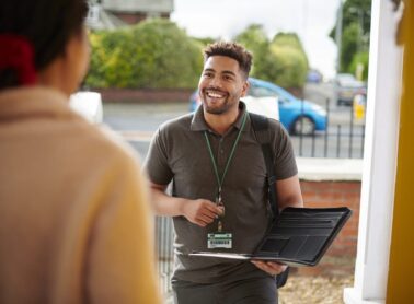 A smiling man with a laptop, ID badge, and backpack stands at a doorstep, actively engaging in resident engagement with another person. Surrounded by greenery and a street in the background, he embodies approachable professionalism outdoors. ©Mobysoft