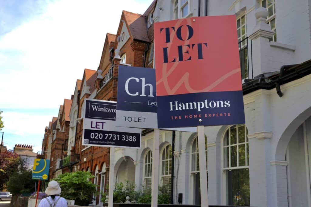 Several "To Let" signs are displayed in front of a row of brick residential buildings. A person wearing a hat and backpack walks past. The scene reflects a residential area with properties available for rent.