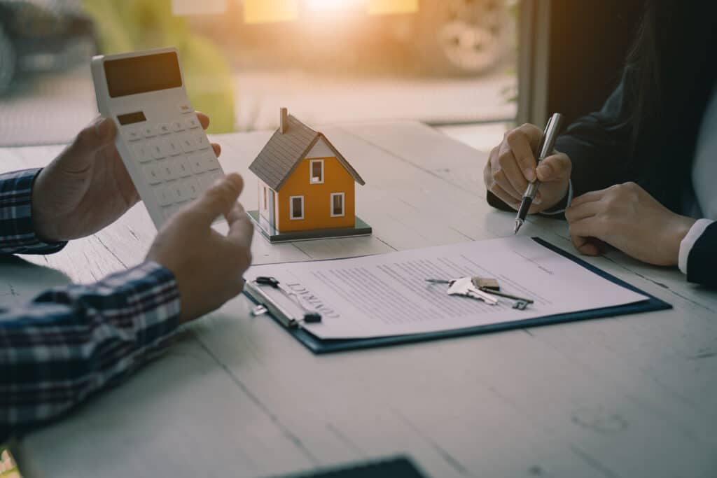 Two people sit at a table discussing real estate. One person holds a calculator, and the other points at a document with a pen. A miniature house model and a set of keys are on the table, suggesting a property transaction. ©Mobysoft