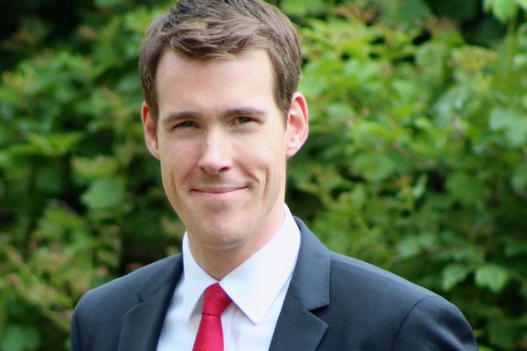 A man in a suit with a red tie smiles while standing in front of a lush green background, as if capturing the spirit of optimism found in social housing stories from the past month. ©Mobysoft