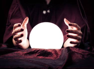 A person in a dark cloak holds their hands around a glowing crystal ball. The scene is dimly lit, casting shadows on the deep red fabric covering the table. The person's nails are painted dark red, as they whisper social housing predictions for 2025, deepening the mysterious atmosphere. ©Mobysoft