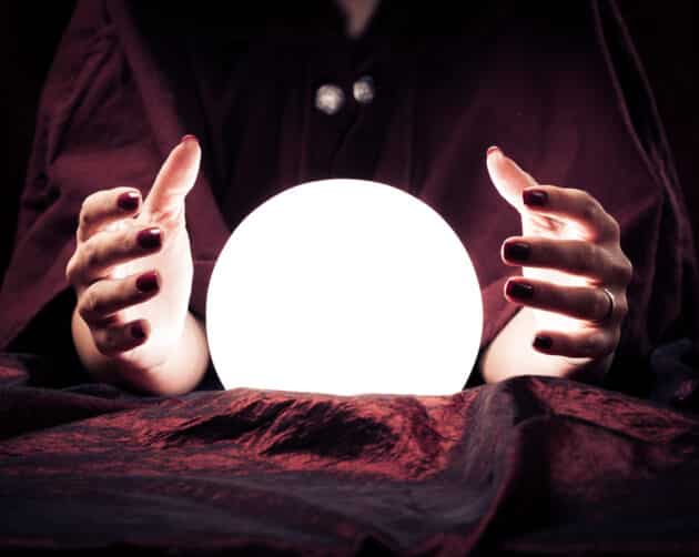 A person in a dark cloak holds their hands around a glowing crystal ball. The scene is dimly lit, casting shadows on the deep red fabric covering the table. The person's nails are painted dark red, as they whisper social housing predictions for 2025, deepening the mysterious atmosphere. ©Mobysoft