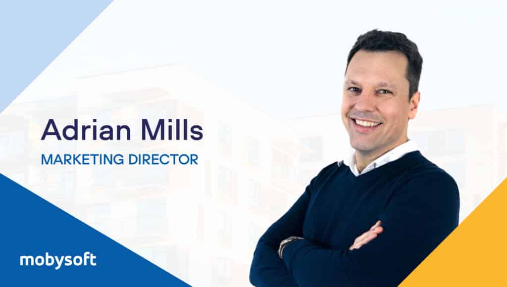 A smiling man with crossed arms stands in front of a blurred building, emphasizing his dedication to social housing. Text reads, "Adrian Mills, Marketing Director." The logo in the bottom left corner says "mobysoft." He is wearing a dark blue sweater over a white shirt. ©Mobysoft