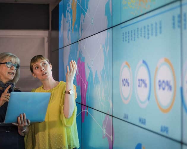 Two women stand in front of a large digital screen displaying graphs and data. One gestures towards the screen while the other holds a laptop, seeming to discuss the information presented. ©Mobysoft