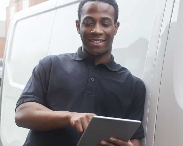 A man in a black polo shirt stands next to a white van, smiling and using a tablet. The background shows a residential area with brick buildings. ©Mobysoft