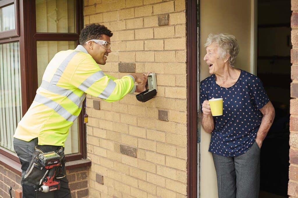 A worker in a high-visibility jacket smiles while installing a device on the brick wall of a house. An elderly woman holding a yellow mug stands at the door, watching and smiling. ©Mobysoft