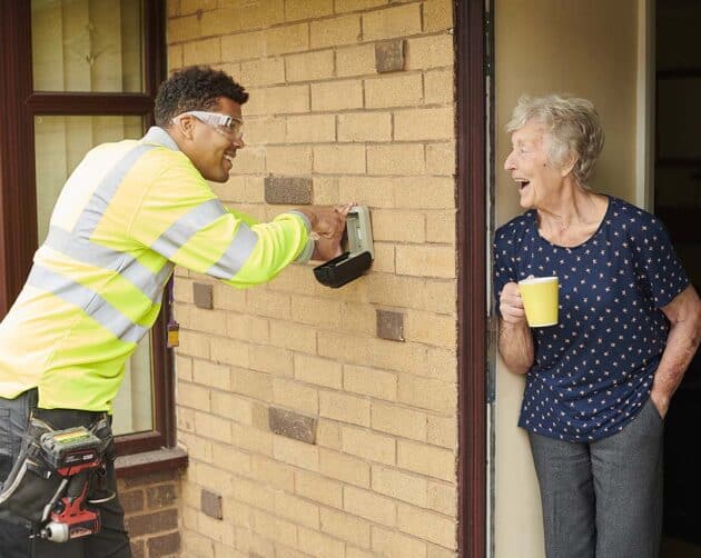 A worker in a high-visibility jacket smiles while installing a device on the brick wall of a house. An elderly woman holding a yellow mug stands at the door, watching and smiling. ©Mobysoft